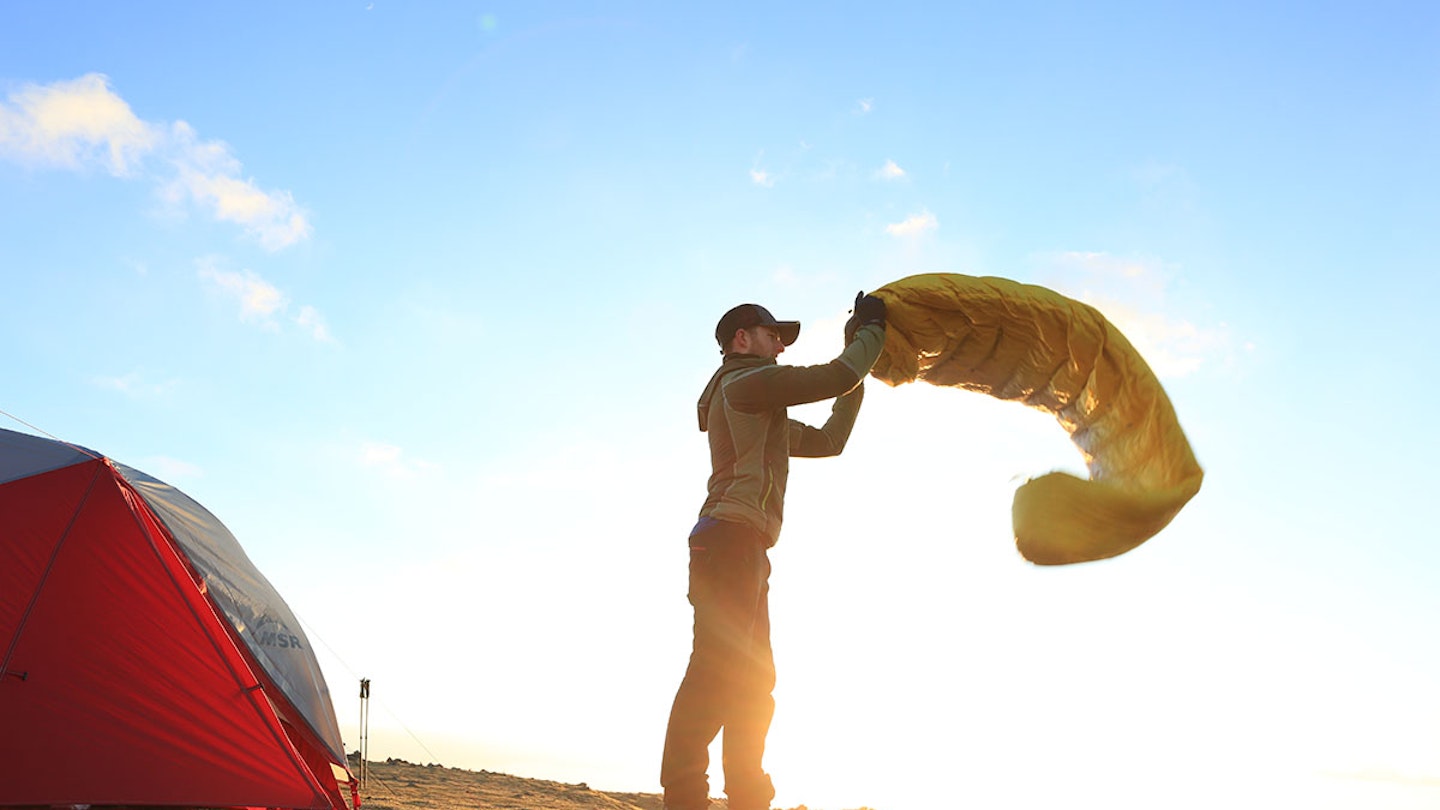 Man shaking out sleeping bag with tent and sky in the mountains